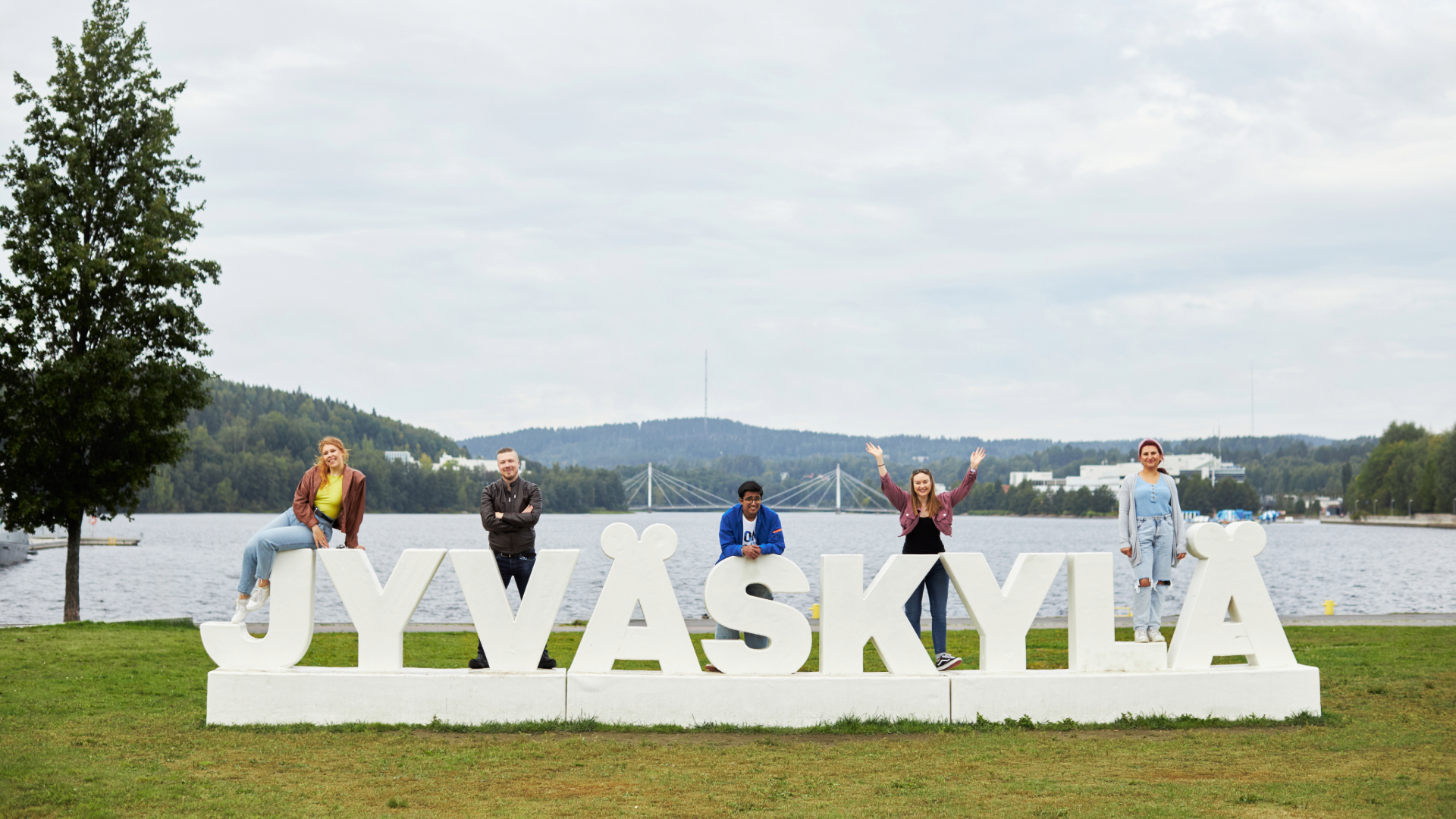 Jyvaskyla written in big letters in grass, with people climbing and smiling on top of them lake and scenery view behind them