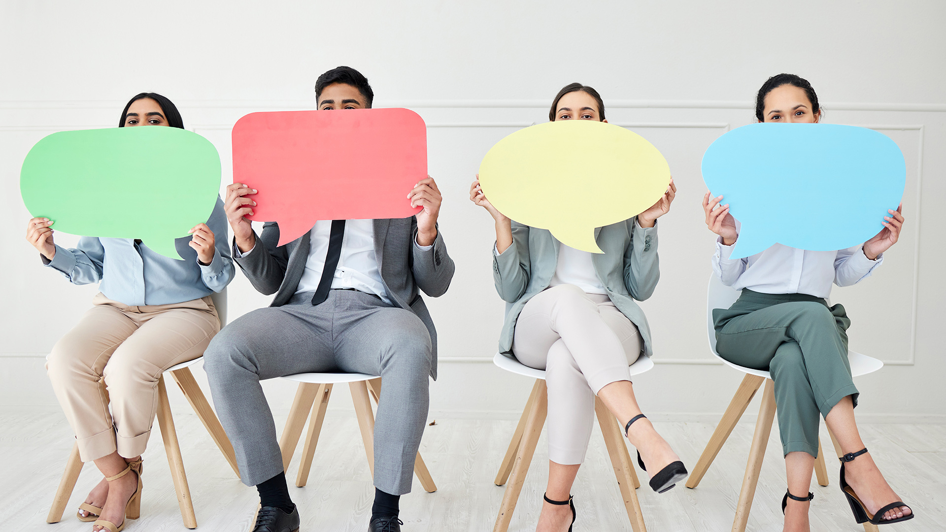 Neljä henkilöä istuu tuoleilla ja kurkistaa edessään olevien puhekuplien takaa.