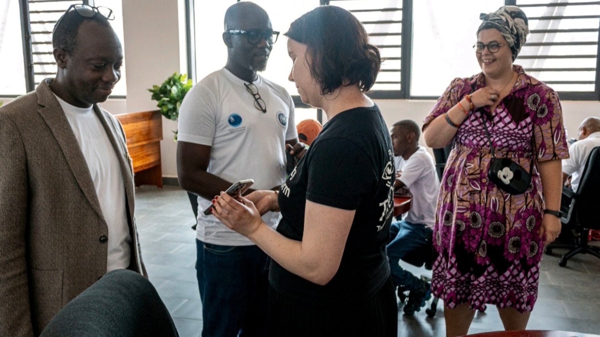 The picture shows four people standing and talking to each other. One of the people is dressed in a colorful purple dress and scarf. One is dressed in a black t-shirt and has dark hair. In the background, you can see light walls, large windows and seated people. One person is wearing glasses on the top of his head and is dressed in a light shirt and a brown jacket. The other person is wearing a white t-shirt and glasses. The atmosphere is relaxed and the event is a workshop.