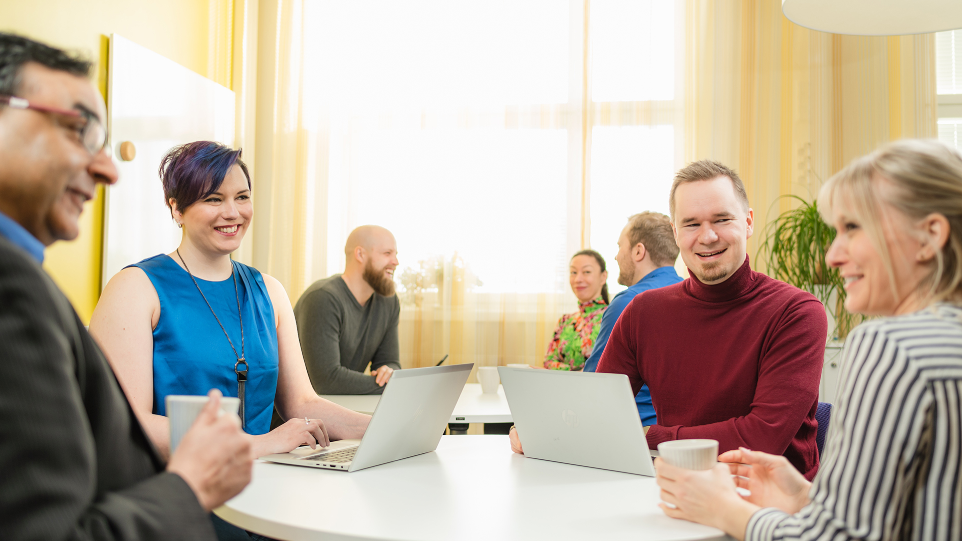 Joukko ihmisiä istuu pöytien ääressä edessään tietokoneita ja käsissään kahvikuppeja.
