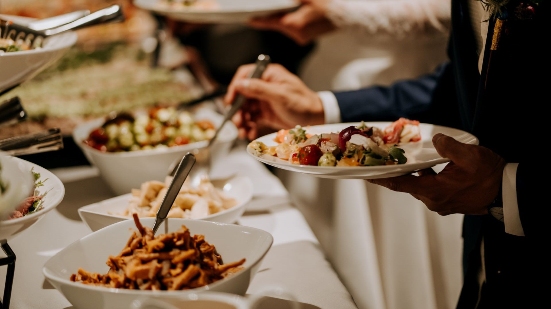 Buffetpöytä, jossa monenlaista ruokaa sekä kaksi henkilöä ottamassa ruokaa lautasilleen.