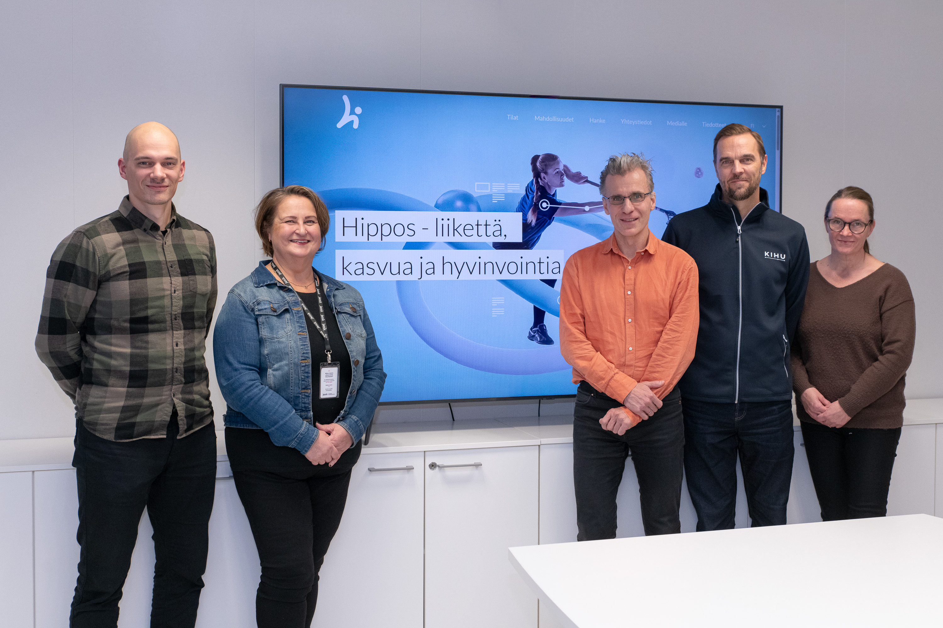 Viisi henkilöä ryhmäkuvassa ison television edessä.