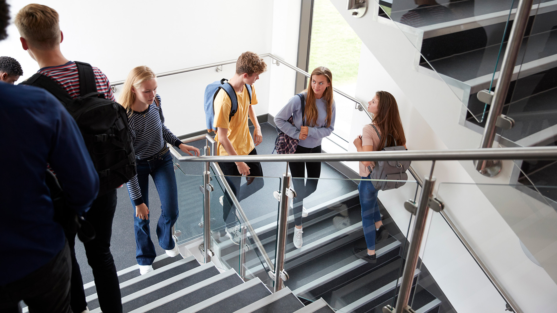 Oppilaitoksen portaikossa ylös ja alas käveleviä nuoria opiskelijoita reput selässään.