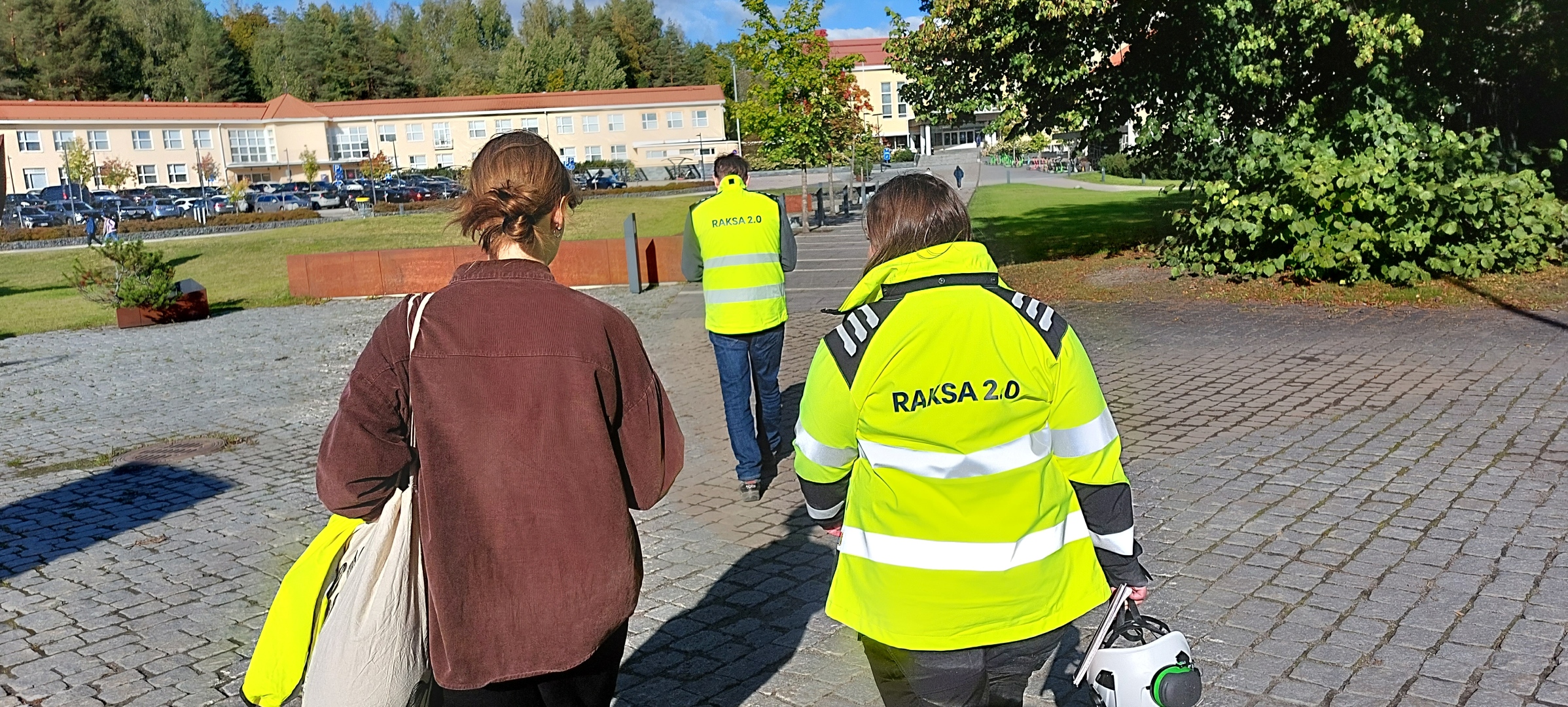 Kolme henkilöä kävelemässä eteenpäin. Kahdella heistä on huomiotakit päällä. Kuvattu ihmisten takaa.