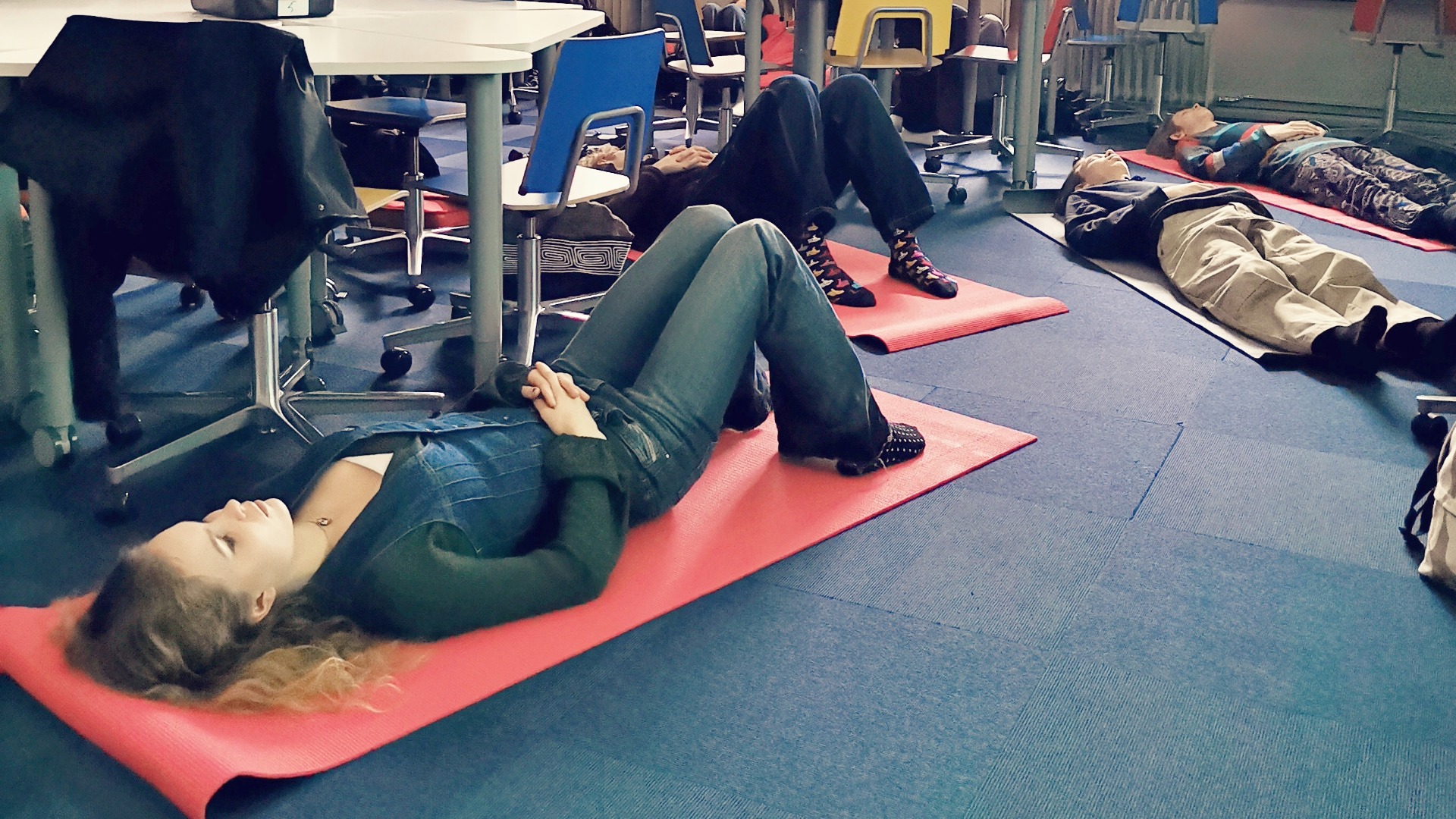 Henkilö makaa selällään joogamatolla silmät kiinni luokkahuoneessa. Taustalla näkyy muitakin oppilaita.