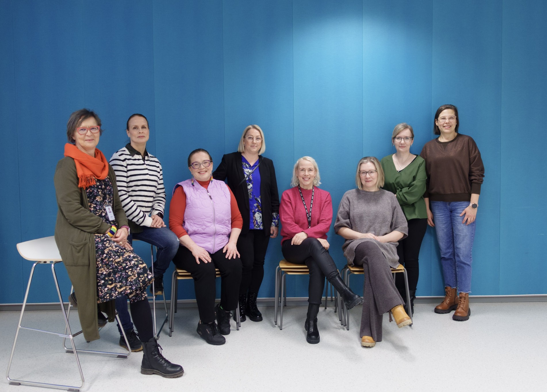 Kahdeksan ihmista on rivissa sinista seinaa vasten