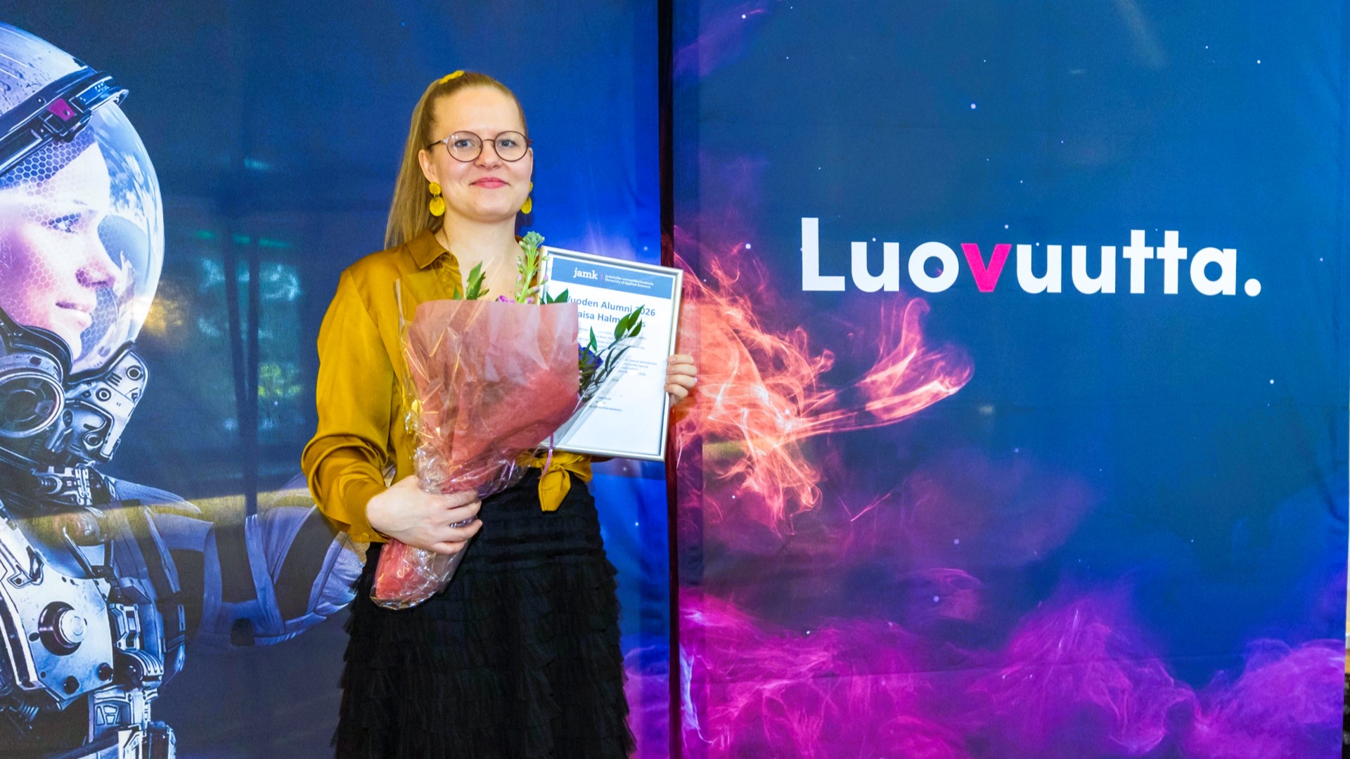 Jamkin Vuoden Alumni Kaisa Halmemies seisoo Jamk-seinäkkeen edessä. Hänellä on kädessään vuoden alumnin kukat ja diplomi.