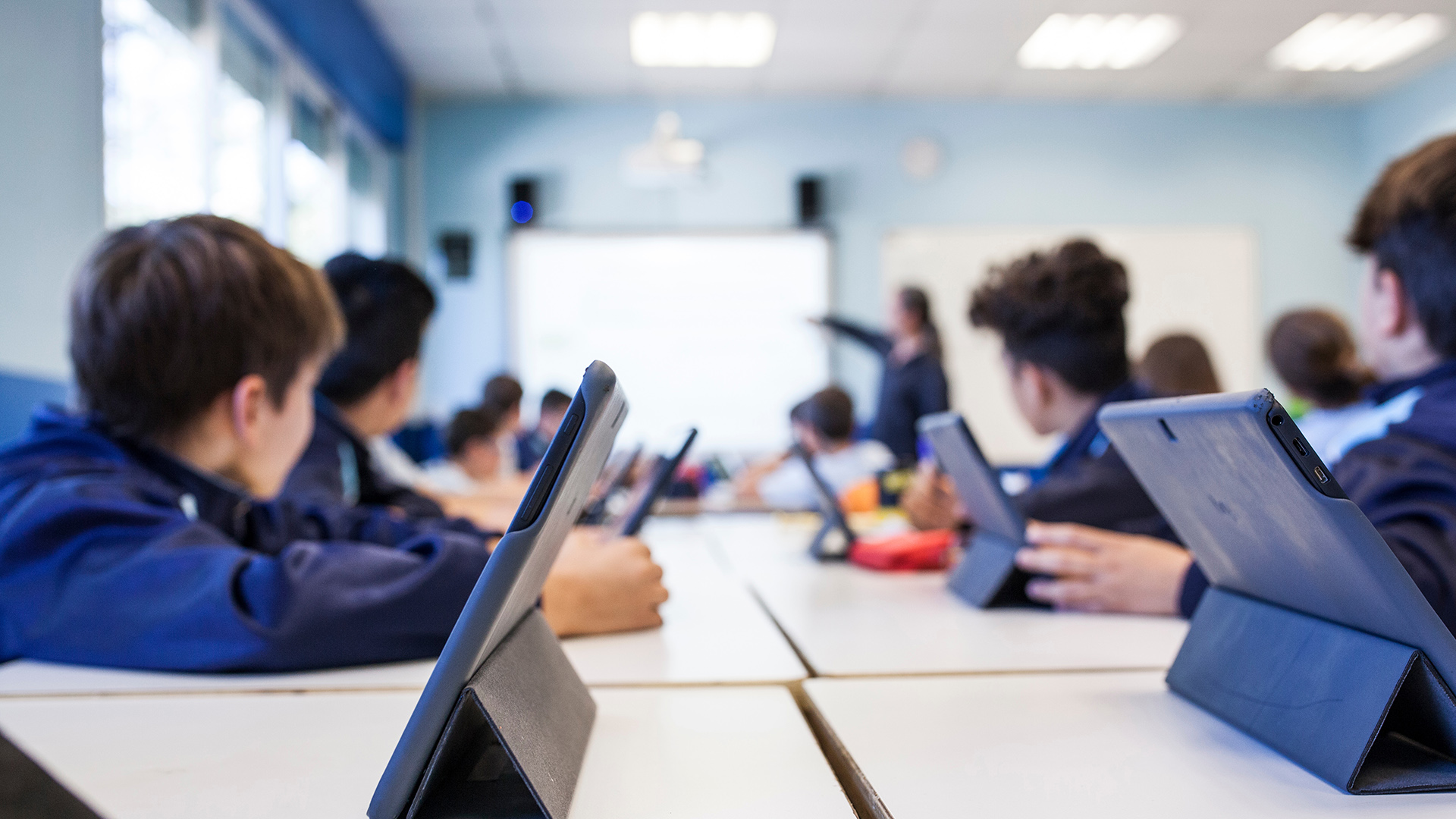 Pöydän ääressä istuvat lapset, joilla on tabletteja, seuraavat taustalla näkyvää opettajaa.