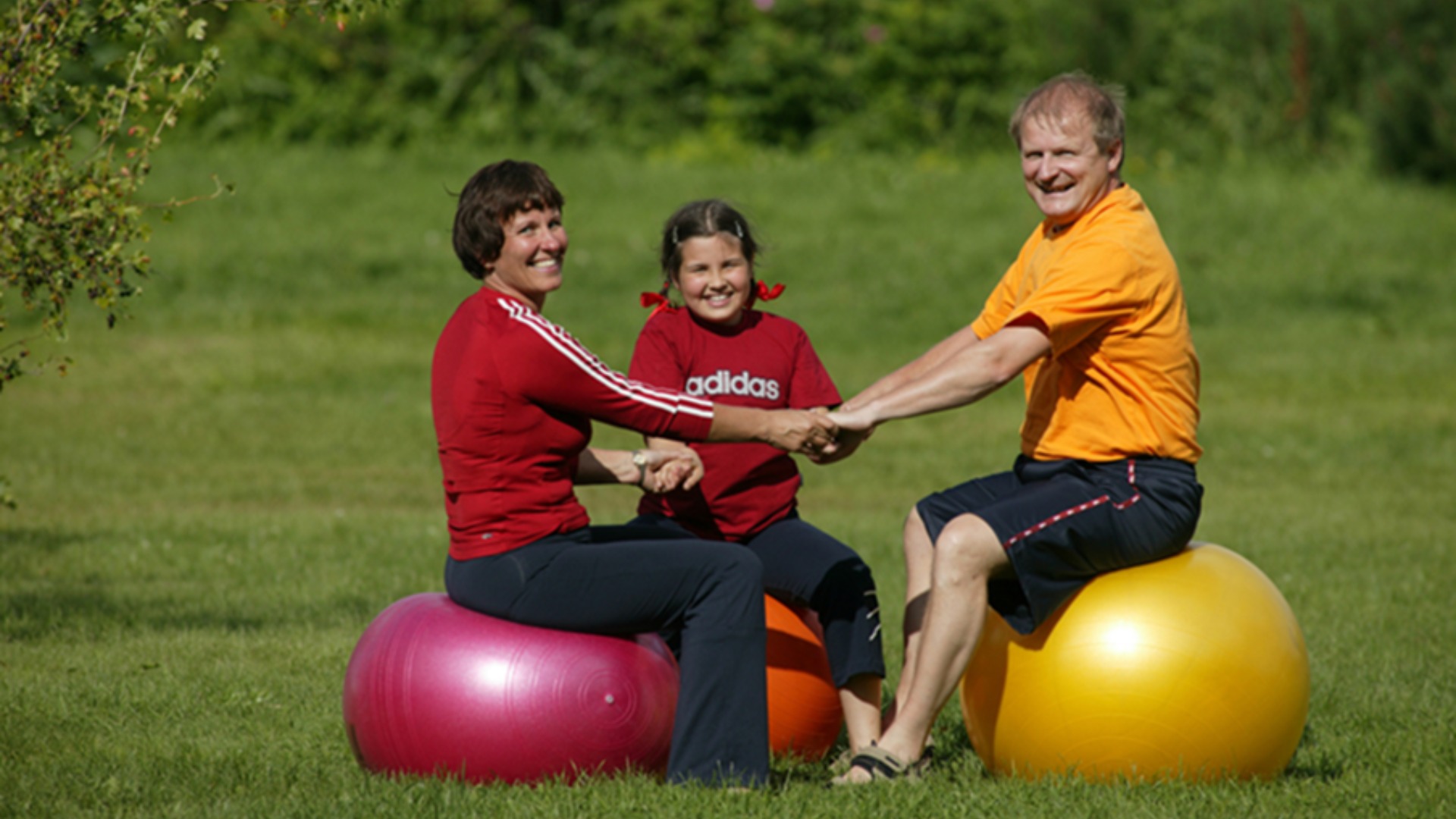 Lapsi, äiti ja isä istuvat jumppapallojen päällä nurmikolla, pitävät toisiaan kädestä kiinni ja katsovat hymyillen kameraan.