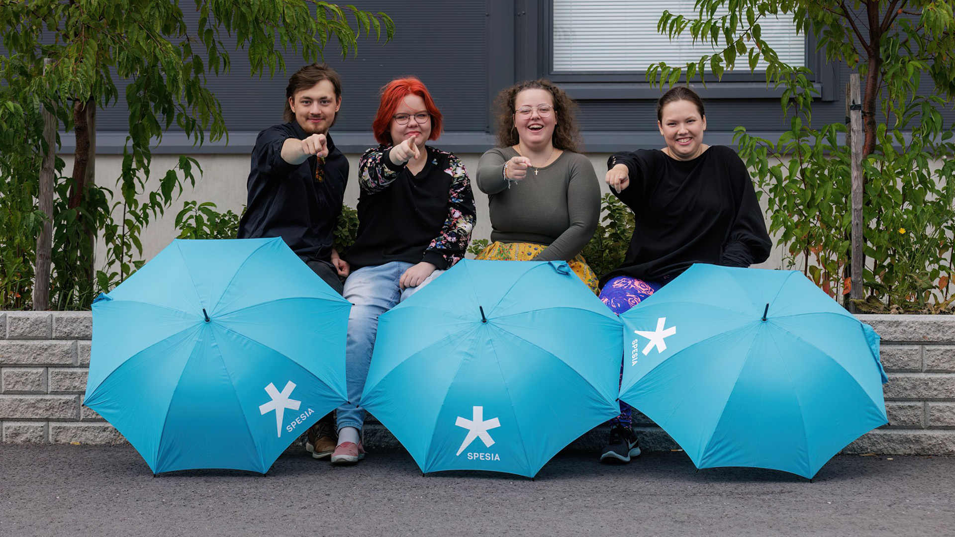 Oppilaitoksen pihassa neljä iloista opiskelijaa istuu ja osoittaa kuvaajaa sormella.