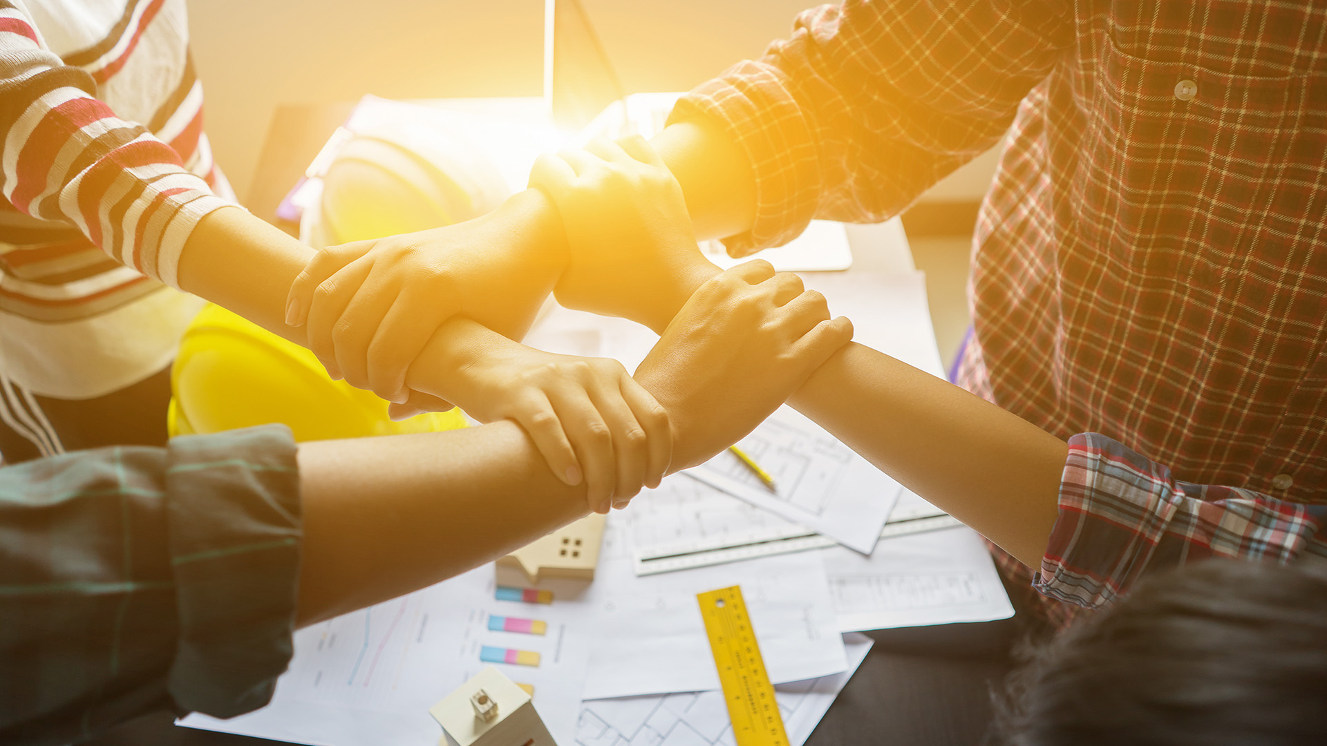 Tiimin kädet yhdistettynä kuvan keskellä, taustalla rakennusalaan liittyviä välineitä ja papereita.