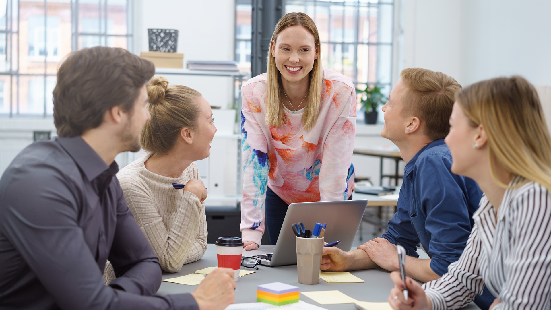 Nuoret opiskelijat työskentelevät ryhmässä ja katsovat iloista opiskelijaa, joka seisoo pöydän päädyssä.