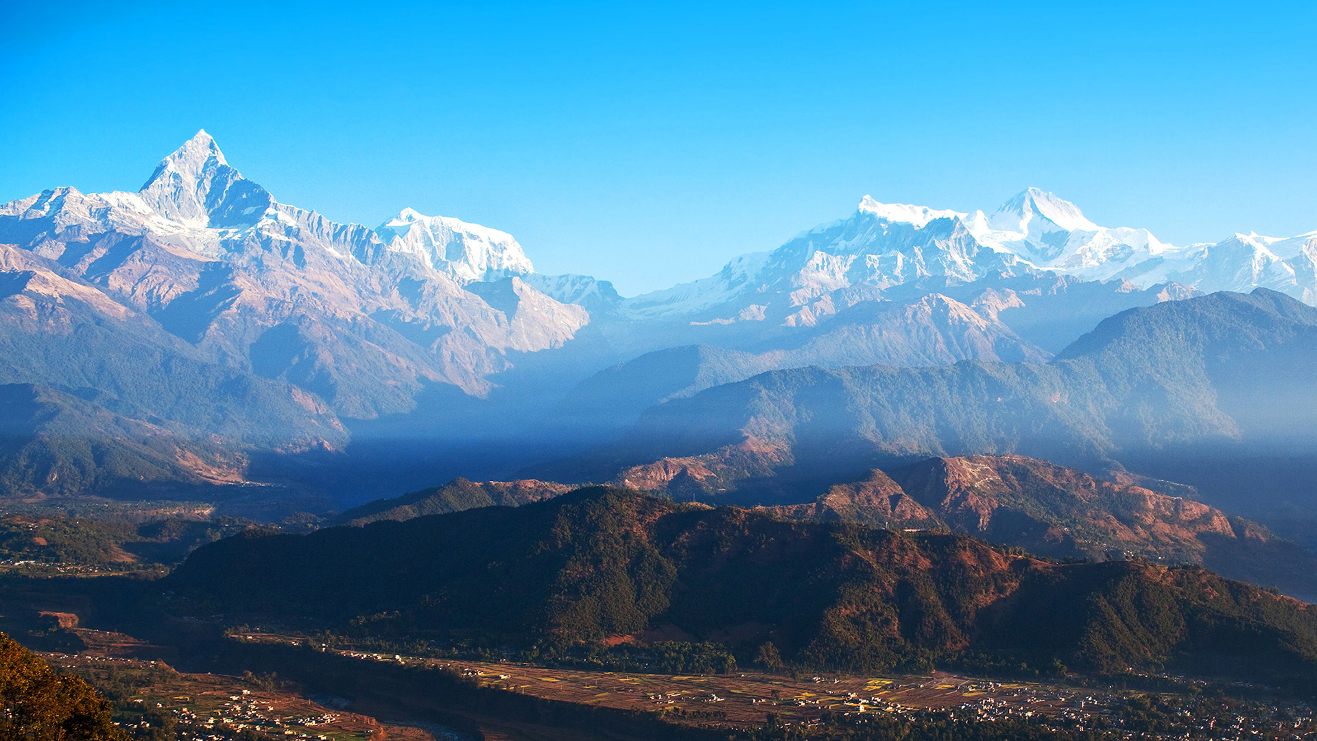 A scenery from Sarangot, Nepal, with mountains and blue sky in the background and villages in front of them.