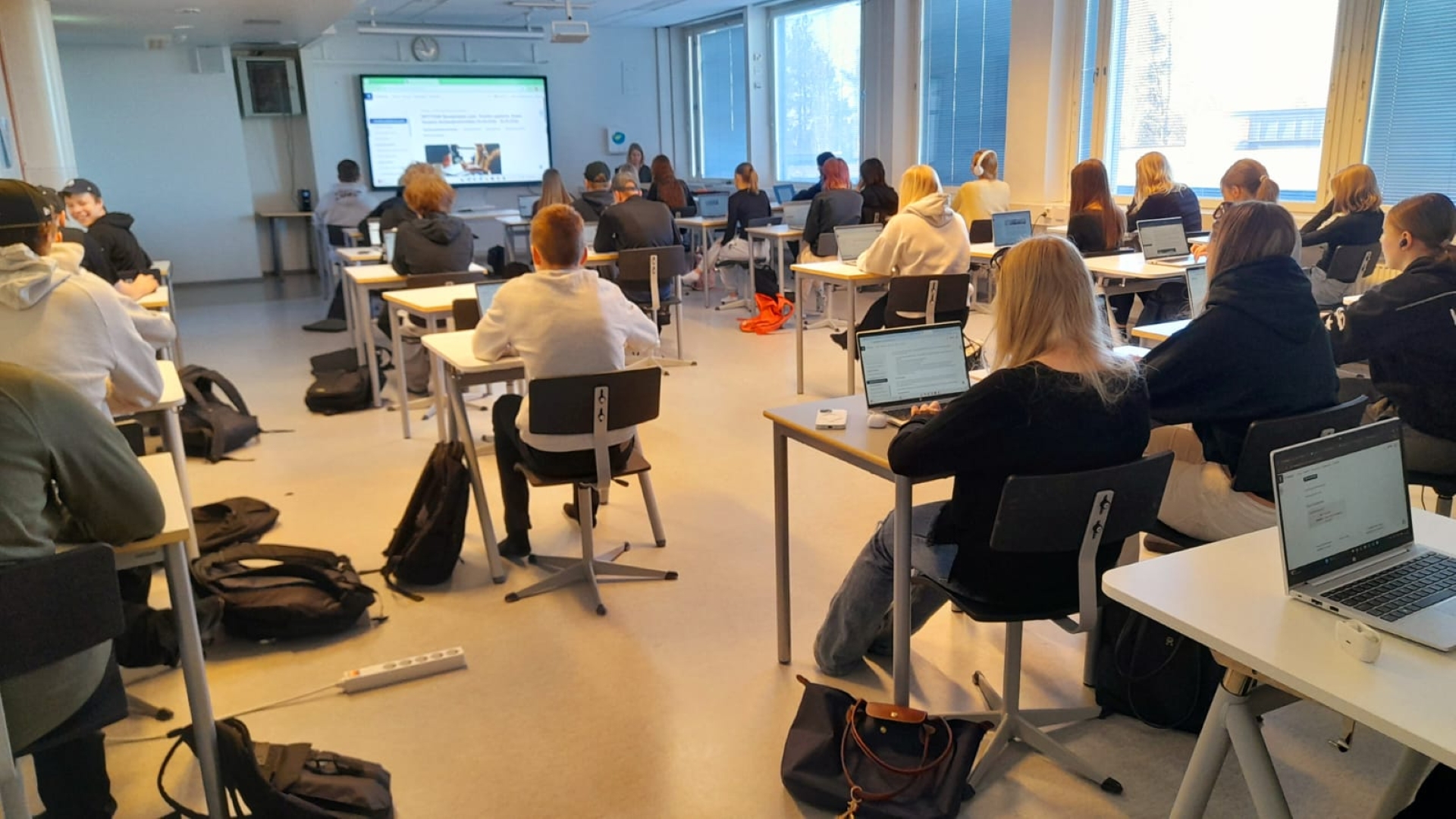 Lukiolaisia opiskeljoita istuu pulpeteissa selin kameraan. Luokan edessä opettaja ohjaa oppilaita Korkeakouluviikon ohjelmaan.