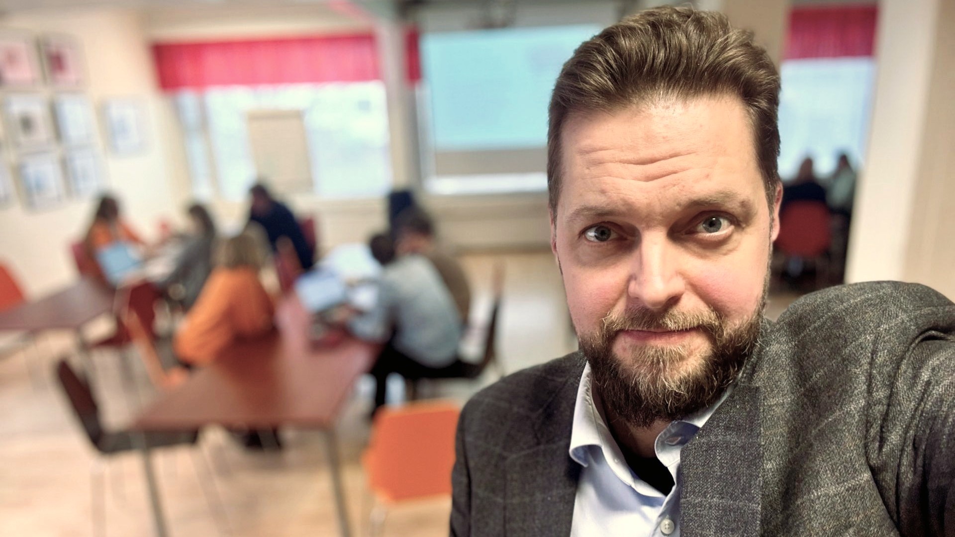 Petri Toikkanen katsoo suoraan kameraan. Hänen takanaan näkyy hämärretysti ihmsiä tekemässä ryhmätöitä.