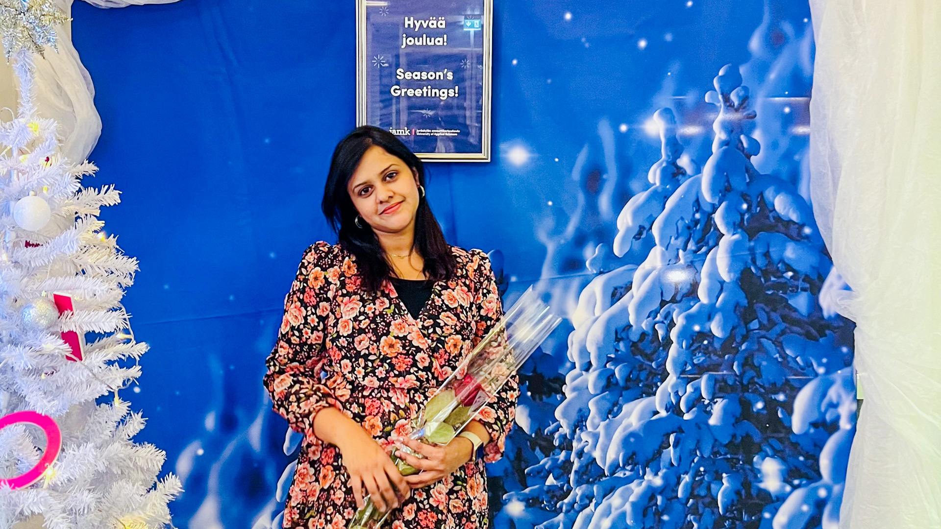 Thisari smiles while holding a rose. A white Christmas tree stands in the foreground, and the text "Season's Greetings!" is visible in the background.