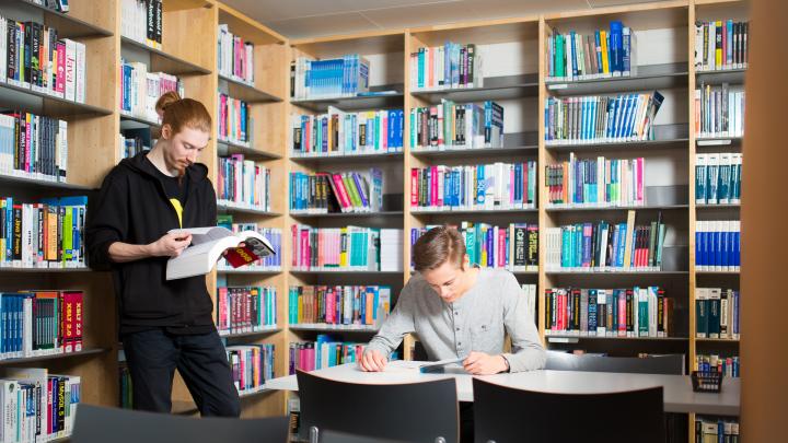 Kaksi opiskelijaa kirjastossa, taustalla kirjahyllyjä