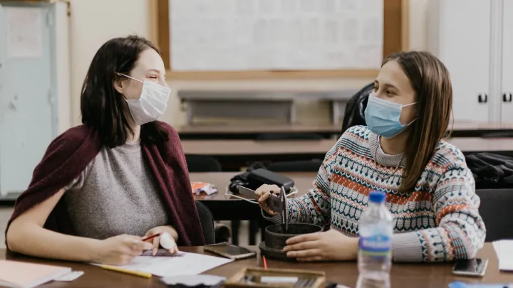 kaksi opiskelijaa katsoo toisiaan pöydän äärellä nenä-suu-suojaimet kasvoillaan 