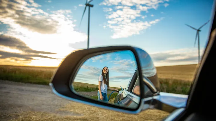 Nainen lataamassa sähköautoa, taustalla tuulivoimala