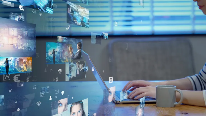 Pöydällä kannettava tietokone ja muki, nuori nainen näppäilee ja edustalla näkyy erilaisia kuvia