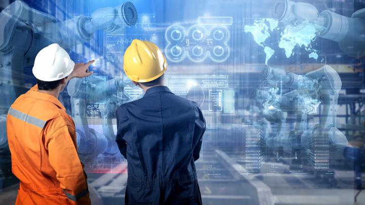 Kaksi työmiestä seisoo haalarit ja kypärät päässä robotiikkatehtassuunnitelmaa katsoen