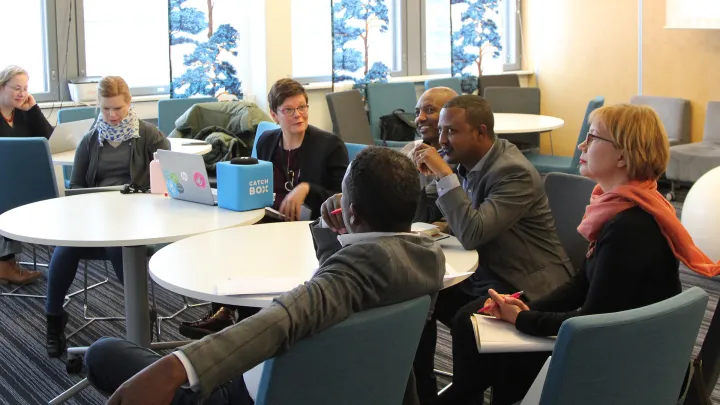 Kuusi henkilöä istuu ja keskustelee pöydän ympärillä. 