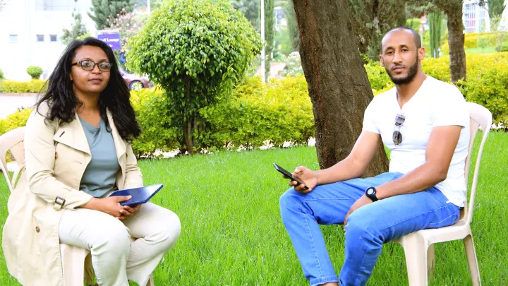 Two people sitting in chairs outside by the school, with a phone and ipad in their hands, grass and green trees around them. 