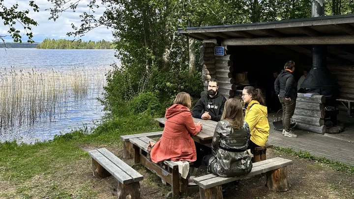 Rannalla pöydän äärellä neljä henkilöä, taustalla hirsinen grillikatos.