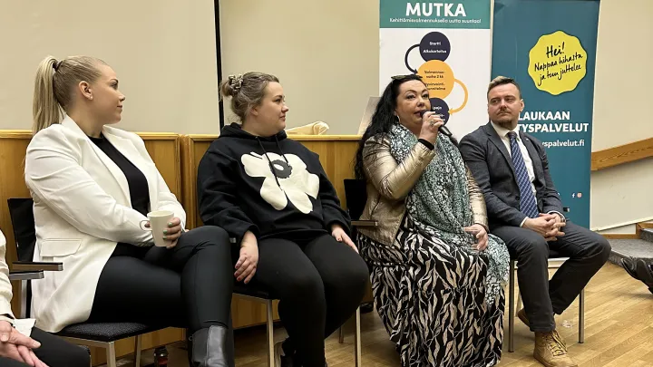 neljä henkilöä auditorion etuosassa istumassa ja yksi henkilö puhuu mikrofoniin