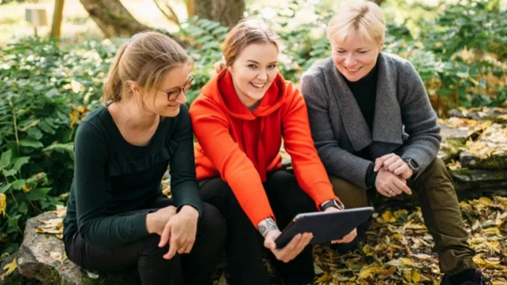 Kolme naista istuvat metsässä ja katsovat ipadia