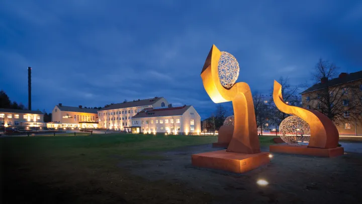 Etualalla metallista tehty suurehko valaistu taideteos, taustalla valaistu rakennus.