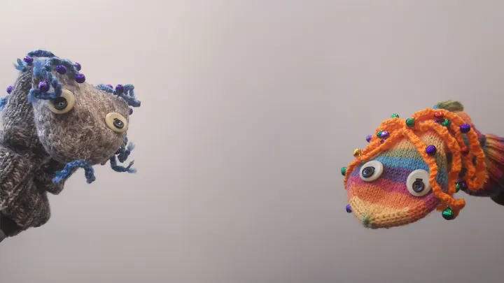 Kaksi itse neulottua sukkamaista käsinukkea, toinen harmaa ja toinen monivärinen.