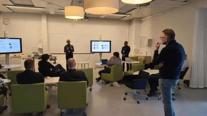 Ihmisiä kokoontuneena luokkatilaan, jossa yksi henkilö on edessä puhumassa muille ja muut kuuntelevat.