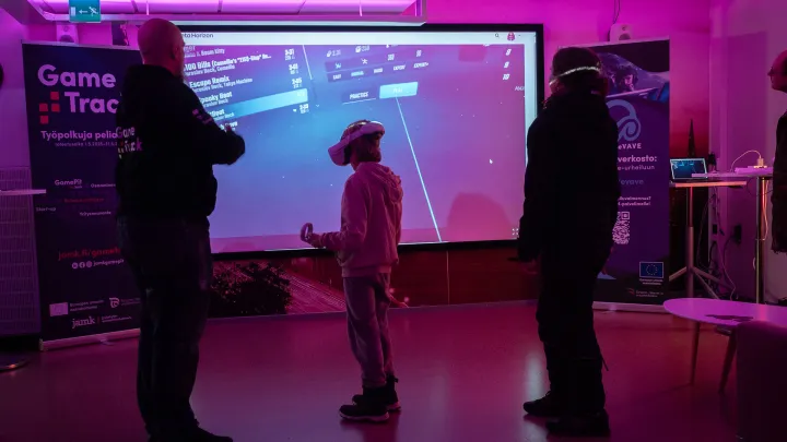 Violettisävyinen kuva, jossa lapsi pelaa VR-laseilla. Kaksi akuista seuraa viereistä.