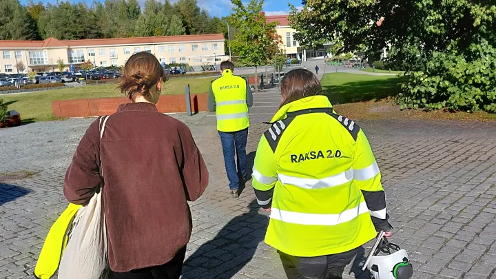 Kolme henkilöä kävelemässä eteenpäin. Kahdella heistä on huomiotakit päällä. Kuvattu ihmisten takaa.