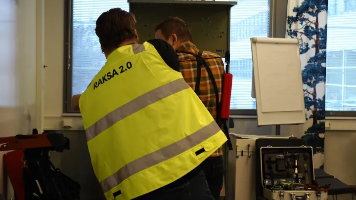Kaksi henkilöä kuvattuna selän takaa. Toisella henkilöllä huomioliivi, jonka selässä lukee RAKSA 2.0. Kuvan henkilöt kokeilevat työvälinettä luokkatilassa.