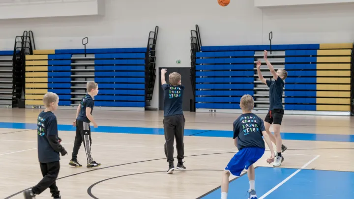 Kuvassa on nuoria pelaamassa koripalloa. Oranssi koripallo on korkealla ilmassa ja yksi nuorista kurkottaa sitä kohti. 