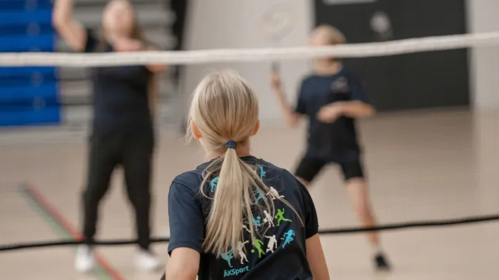 Kuvassa nuori pelaamassa sulkapalloa. Verkon toisella puolella on kaksi nuorta. Kaikilla on päällään tummansiniset harjoittelupaidat, jonka selässä on kuvakkeita urheilulajeista ja teksti ÄKSport.