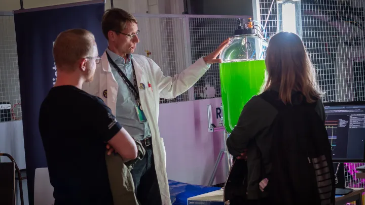 Valkotakkinen mies esittelee laboratorioympäristössä kahdelle selin kameraa olevalle henkilölle lasista suurta astiaa, jonka sisällä on kirkkaan vihreää lientä, mikrolevää.